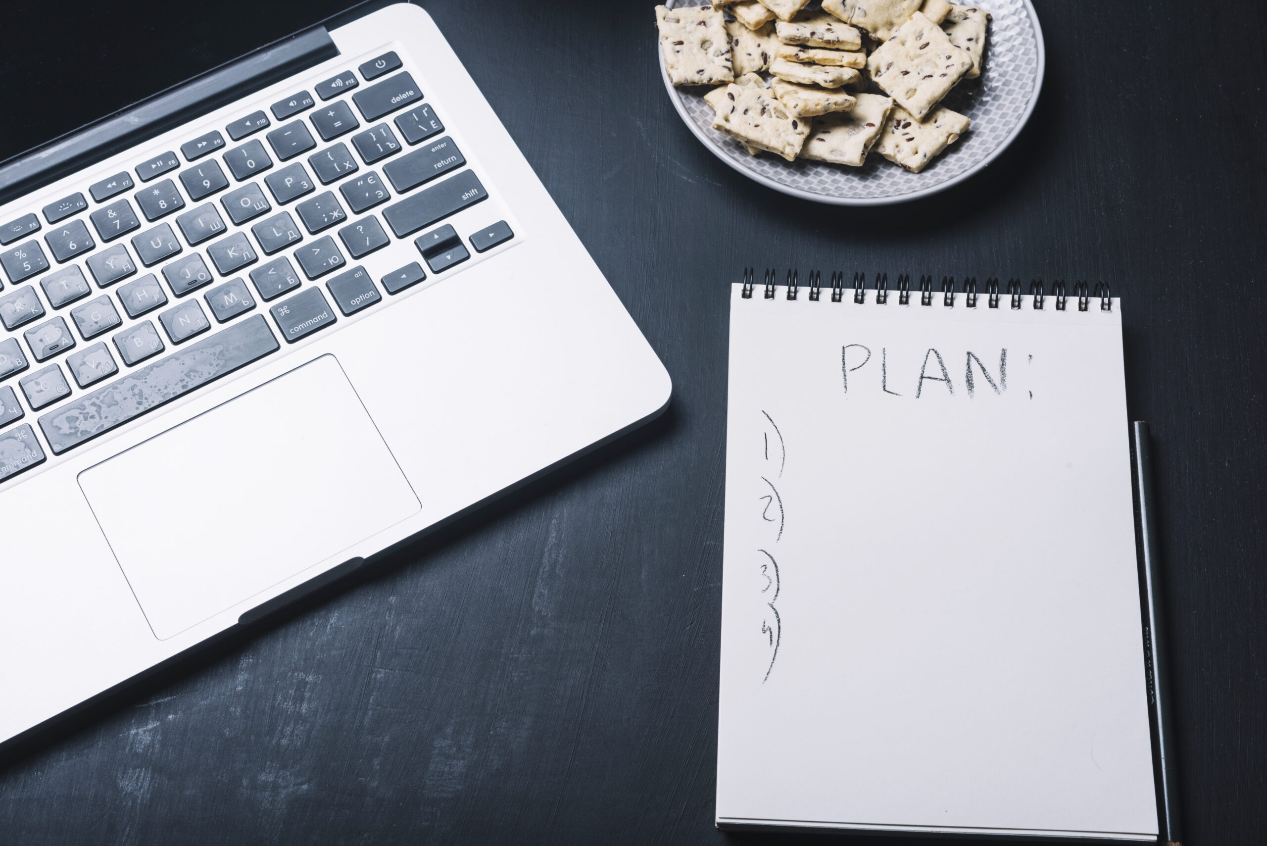 notepad-laptop-concept Spiral notepad with the word PLAN written and numbered lines beside a silver laptop on a dark desk